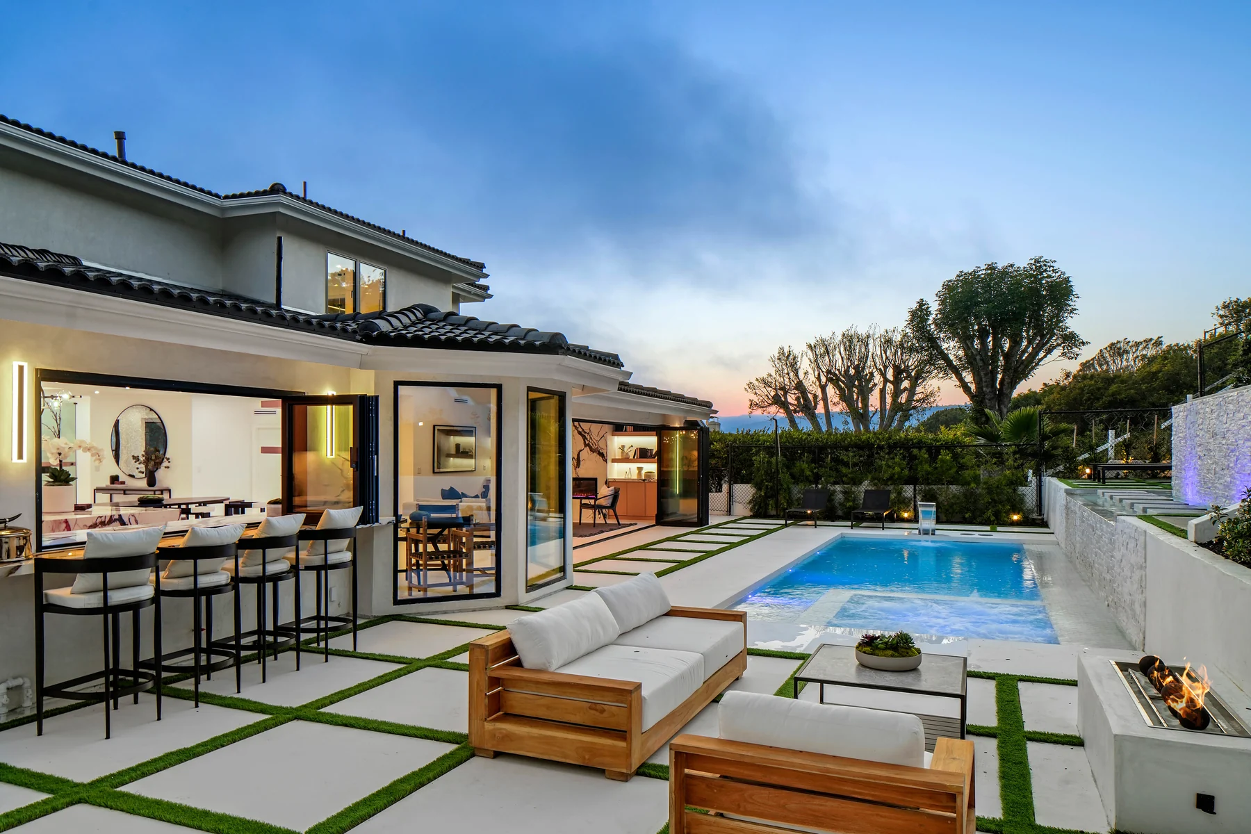 Wide rear elevation at dusk, glass walls open onto the pool terrace with the interior lit behind