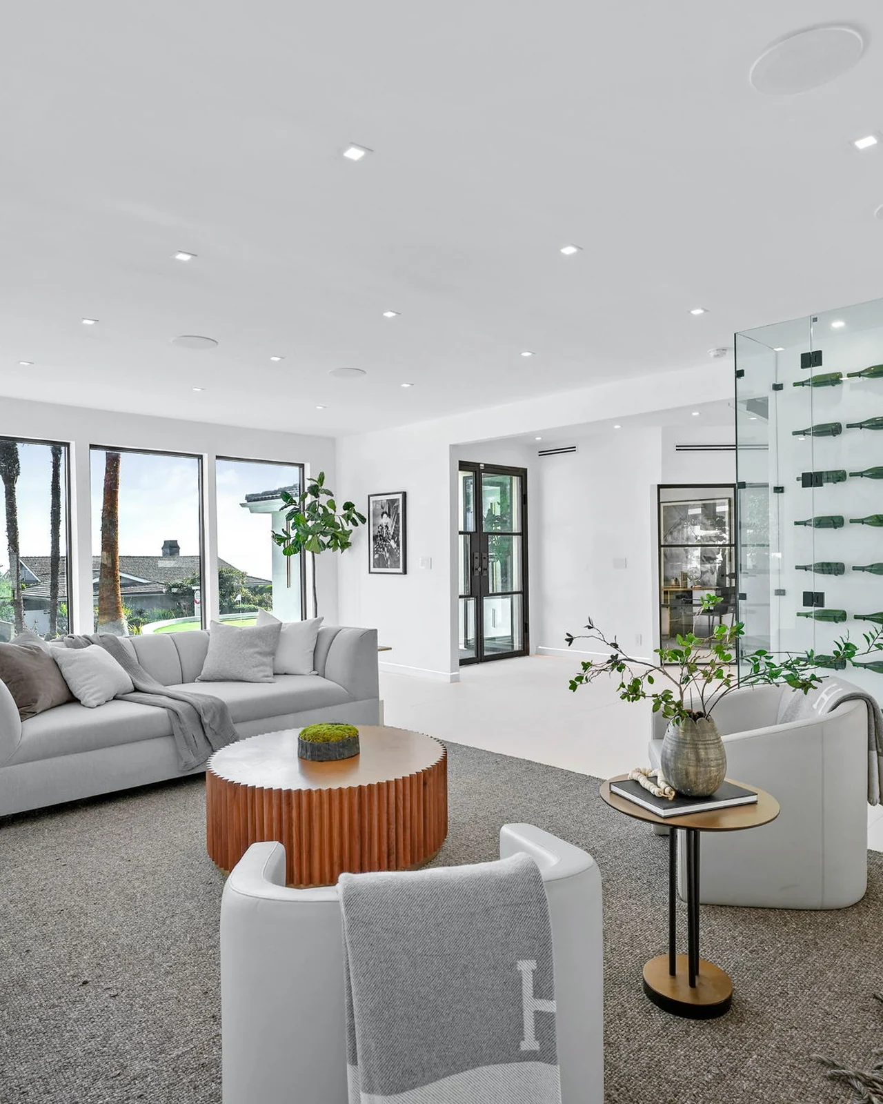 The same zone rebuilt with a pale seamless floor, curved cream lounge chairs, a drum coffee table and a lit glass-shelved wine display wall